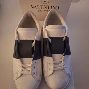 Valentino Garavani skor - Säljer nu ett par snygga vita sneakers från Valentino Garavani med bred marinblå rem över vristen och coola nitar på sulan. Skorna har rund tå, platt sula och klassisk snörning. Tillverkade i skinn med lyxig känsla och kommer med originalkartong och dustbag.