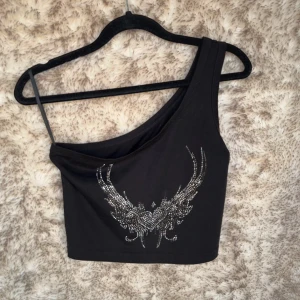 Svart one shoulder topp med strass - Svart croppad one shoulder topp med glittrande strassdetaljer i form av vingar och hjärta framtill.