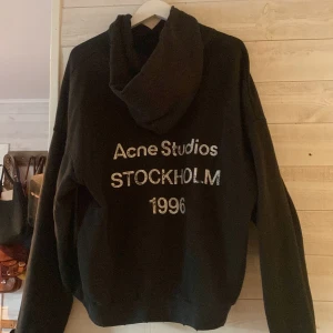 Svart hoodie Acne Studios Stockholm - Svart hoodie från Acne Studios med tryck i vitt på ryggen: 'Acne Studios STOCKHOLM 1996'. Klassisk huva och långärmad modell, tillverkad i mjukt bomullsmaterial. Perfekt för dig som gillar stilrena streetwear-plagg med tydlig logga. Storlek S