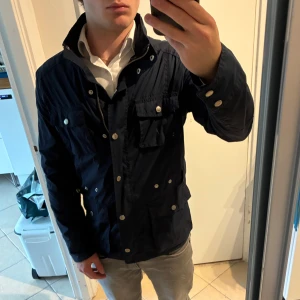 Mörkblå fältjacka från Boomerang - Snygg mörkblå vindjacka från Boomerang med flera fickor och silverfärgade knappar. Jackan har dragkedja framtill, hög krage och långärmade ärmar. Perfekt för dig som gillar stilren och funktionell design. Materialet är lätt och syntetiskt. Bra skick. Storlek 40, sitter som S