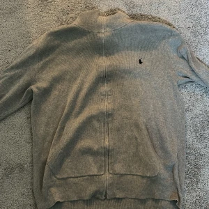 Grå kofta från Polo Ralph Lauren - Snygg grå kofta från Polo Ralph Lauren med dragkedja framtill och diskret logga broderad på bröstet. Tröjan är långärmad och har ribbade muddar vid ärmslut och nederkant. Perfekt för dig som gillar klassisk stil med en modern touch. Det ända dåliga med den har den har igen sytt hål uppe vid kragen men det är inget som syns när den är ner fälld 