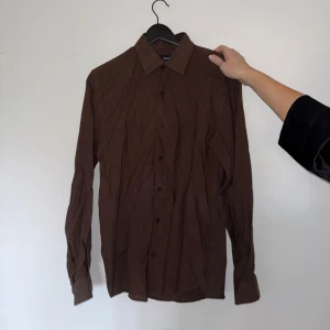 Brun skjorta från Antioch, regular fit - Brun långärmad skjorta från Antioch i regular fit. Skjortan har klassisk krage, knäppning framtill och vid ärmslut samt är tillverkad i ett mjukt bomullstyg. Perfekt för dig som gillar en enkel och stilren look.