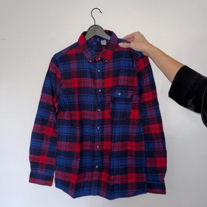 Rutig skjorta från H&M Divided - Röd och blå rutig skjorta från H&M Divided i regular fit. Skjortan har klassisk krage, bröstficka med knapp och långa ärmar med manschettknappar. Tillverkad i mjuk bomull, perfekt för en chill och avslappnad stil.