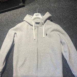 Grå hoodie från Polo Ralph Lauren - Snygg ljusgrå hoodie från Polo Ralph Lauren med dragkedja framtill och klassisk logga broderad på bröstet. Tröjan har huva med snörning och två fickor framtill. Perfekt för chill dagar eller när du vill ha en clean look.