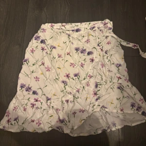 Blommig omlottkjol från Vila - Supersöt kort omlottkjol från Vila i vitt med blommigt mönster i lila, grönt och gult. Kjolen har volangkant och knytband i midjan. Aldrig använd🌸