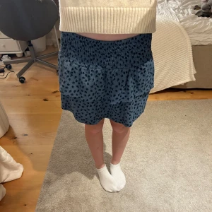 Blå volangkjol med svarta prickar - Supersöt blå kortkjol med svarta prickar och volangdetaljer. Kjolen har smock i midjan för en bekväm passform och är gjord i ett mjukt bomullstyg. Perfekt för dig som gillar en lekfull och trendig stil. Kjolen är lite nopprig men inget som syns på långt håll. Kjolen är i storlek 152 men är ganska stor i storlek. Själv är jag 159 cm och bär vandligtvis 158 eller XS och den är absolut inte för liten.