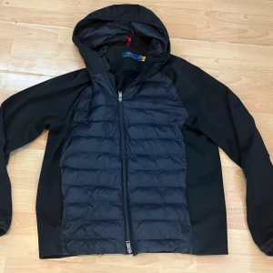 Svart Hybridjacka från Polo Ralph Lauren - Säljer nu min Svart Hybridjacka från Polo Ralph Lauren med huva som sitter som L/M. Jackan har quiltad front, släta ärmar och sidopaneler i syntetmaterial. Insidan har en praktisk ficka med dragkedja. Perfekt för kalla dagar och snygg streetstil. Jackan är använd ett fåtal gånger.