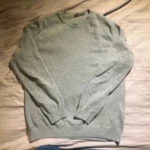 Grå ulltröja från Oscar Jacobson - Klassisk grå rundhalsad tröja från Oscar Jacobson i mjuk ullmix. Tröjan har ribbade muddar vid ärmslut, hals och nederkant. Perfekt för lager-på-lager och enkel att matcha med jeans eller chinos.