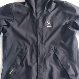 Svart regnjacka från Haglöfs - Snygg svart regnjacka från Haglöfs med huva och dragkedja framtill. Jackan har två snedställda fickor och diskret logga på bröstet. Tillverkad i slitstarkt material och märkt med 'Proof' och 'Climatic' för extra väderskydd.