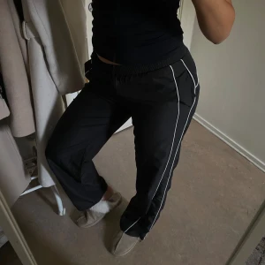 Svarta träningsbyxor med vita detaljer - Svarta träningsbyxor med vita ränder längs sidorna. Byxorna har en loose passform och är tillverkade i ett syntetmaterial som känns lätt och smidigt. Perfekta för en sportig look eller chill dagar. Byxorna är helt nya🫶🏼