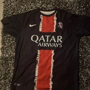 PSG fotbollströja Nike blå/röd - Paris Saint-Germain fotbollströja från Nike i mörkblått med röd vertikal rand och vita detaljer. Tryck med Qatar Airways på bröstet och PSG-logga på vänster sida. Klassisk passform och kortärmad modell, perfekt för fotbollsälskare.