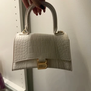 Vit handväska  - Stilren vit handväska från  med krokodilpräglat mönster och guldfärgad B-detalj framtill. Väskan har ett stadigt handtag och är tillverkad i skinnliknande material. Perfekt för dig som vill ha en trendig och lyxig accessoar.