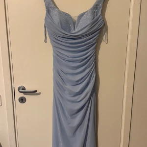 Ljusblå långklänning med strass - Säljer en ljusblå långklänning med glittriga strassdetaljer på axelbanden och över bysten. Klänningen har draperad design framtill och smickrande passform. Perfekt för bal eller festliga tillfällen. Materialet känns mjukt och följsamt.