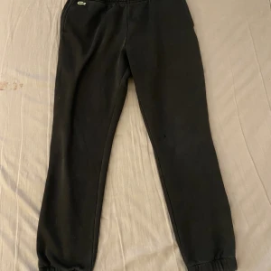 Lacoste  - Svarta mjukisbyxor från Lacoste med klassisk grön krokodillogga på höften. Byxorna har elastisk midja och muddar vid bensluten för en skön passform. Perfekta för chill eller träning, tillverkade i mjukt bomullsmaterial.