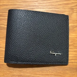 Svart skinnplånbok från Ferragamo - Stilren svart plånbok från Ferragamo i exklusivt präglat skinn. Plånboken har flera kortfack, ett fönster för ID och diskret Ferragamo-logo i silver på framsidan. Perfekt för dig som vill ha en klassisk och lyxig accessoar i fickan.