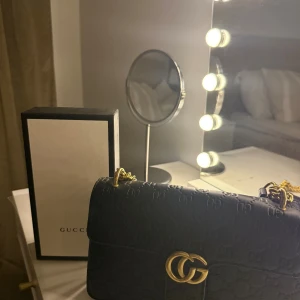 Svart axelväska  - Säljer en svart axelväska  i skinn med GG-logga i guld framtill och präglat mönster över hela väskan. Väskan har en snygg guldfärgad kedjerem och klassisk rektangulär form. Perfekt för dig som vill ha en lyxig och trendig accessoar.