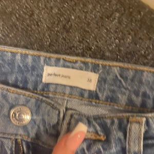 Blå jeans från Perfect Jeans, storlek 36 - Snygga blå jeans från Perfect Jeans i storlek 36. Modellen har klassisk low waist, slitningar och hål på ena benet för en trendig look. Jeansen är bootcut och är tillverkade i ett mjukt bomullsmaterial med dragkedja och knapp framtill.