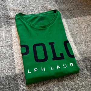 Grön Polo Ralph Lauren t-shirt - Snygg grön t-shirt från Polo Ralph Lauren med stor svart logotyp på bröstet och vit text under. Klassisk rund hals och korta ärmar. Tillverkad i mjuk bomull som är skön mot huden. Perfekt för dig som gillar stilrena och sportiga plagg.