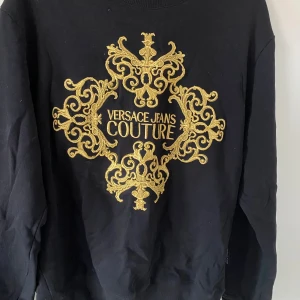 Svart sweatshirt Versace Jeans Couture - Svart sweatshirt från Versace Jeans Couture med stort broderat guldigt barockmönster och logga på bröstet. Tröjan har rund halsringning och långa ärmar. Perfekt statement-plagg för dig som gillar lyxig streetstyle.