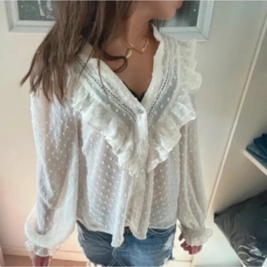 Vit volangblus med prickar från Zara - Supersöt vit blus från Zara med volangdetaljer längs v-ringningen och ärmsluten. Prickigt mönster i tyget, knappar framtill och luftiga, långa ärmar. Perfekt för dig som gillar romantisk och trendig stil. Ena armen är lösare än den andra, men inget man märker