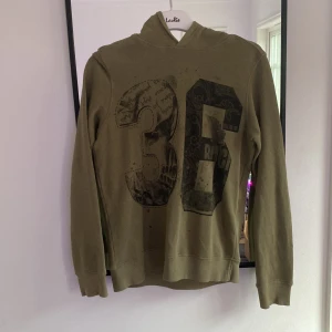 Olivgrön hoodie med tryck från Sisley - Olivgrön hoodie från Sisley med stort svart tryck av siffran 36 på framsidan. Tröjan har huva, långa ärmar och är gjord i mjuk bomull. Perfekt för dig som gillar streetstyle och vill ha en chill look.