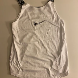 Vit linnetopp från Nike med logoband - Säljer en vit linnetopp från Nike med svarta axelband där det står 'NIKE' i vitt. Toppen har en svart Nike-logga framtill och är tillverkad väldigt mjuk. Perfekt för en sport. BARN STORLEK