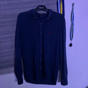 Blå långärmad skjorta från Ralph Lauren - Snygg blå långärmad skjorta från Ralph Lauren med klassisk krage och knappar framtill. Skjortan har det ikoniska röda Polo-logot på bröstet och är tillverkad i mjuk bomull. Perfekt för dig som gillar stilrena och tidlösa plagg. Färgen är något sliten men vid användning så är det inget man tänker på, tveka inte på att höra av dig vid minsta lilla fundering😁 priset är ej hugget i sten🤩