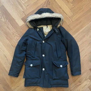 Svart parkasjacka från Woolrich - Säljer en svart parkas jacka från Woolrich med huva med päls. Jackan är workwear fårn Alaska och garanterar att du är varm i vinter. Det finns knappar för att stänga och dragkedja. Stora fickor och värme fickor. Den är storlek S EU USA XS men skulle personligen säga den passar S mer  upp mot M. 