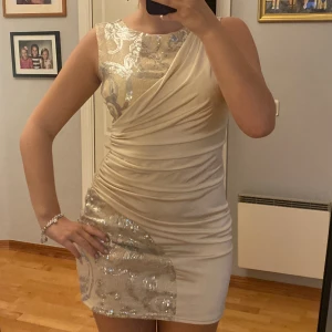 Champagnefärgad kort klänning med paljetter - Säljer en champagnefärgad kort klänning med ett glittrigt paljettmönster i silver på axelpartiet och nedre delen. Klänningen har omlott-detaljer, är ärmlös och sitter tight med snygg rynkning längs sidorna. 