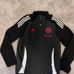 Manchester United vindjacka Adidas - Svart vindjacka från Adidas med Manchester United-logga och Tezos-tryck i rött på bröstet. Jackan har vita ränder på axlarna, hel dragkedja framtill och långärmad design. Perfekt för fotbollsfans som vill ha en sportig look.