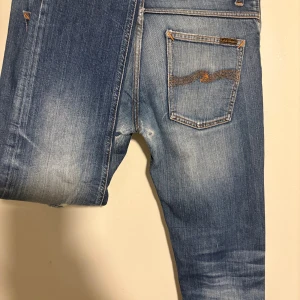 Nudie Jeans blå slitna jeans - Blå jeans från Nudie Jeans med slitningar och hål på framsidan samt bak vid grenen. Klassisk femficksmodell med orange sömmar och broderad bakficka. Jeansen har en ljus tvätt och är tillverkade i bomull. RIKTIGT snygga och stilrena . W30 L31 Modell: grim tim 