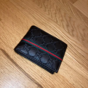 Svart/Grön/Röd plånbok i skinn - Snygg svart plånbok i äkta skinn med präglade GG-loggor och klassisk grön-röd rand på framsidan. Plånboken har flera kortfack och plats för sedlar. Perfekt accessoar för dig som gillar lyxiga detaljer. 