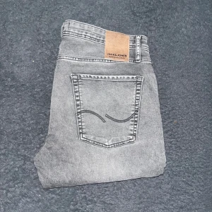 Jack & Jones jeans  - Snygga grå jeans från Jack & Jones med slim fit och klassisk femficksdesign. Jeansen har en ljusgrå tvätt med subtila slitningar och svarta sömmar. Tillverkade i stretchig denim för en bekväm passform. Perfekta för dig som gillar en modern och smal siluett. Midja 42cm, längd 105cm. JAG SKICKAR INGA FLER BILDER NÄR JAG BÄR JEANSEN :)