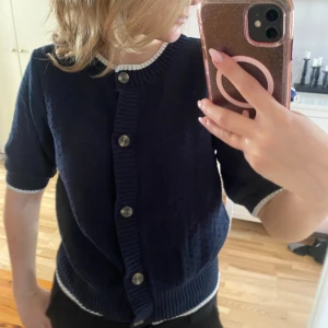 Holly&Whyte kofta - Snygg marinblå kofta från Holly & Whyte med kort ärm och vita ribbade kanter. Koftan har knappar framtill och ett diskret rutmönster i stickningen. Perfekt till lager-på-lager och funkar året runt.