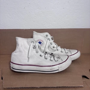 Vita Converse All Star high-top sneakers - Klassiska vita Converse All Star high-top sneakers med snörning och rund tå. Skorna är i canvas med vit gummisula och röda samt svarta detaljer runt sulan. Ikonisk Converse-logga på sidan och bak på hälen. Perfekta för en avslappnad och cool stil.