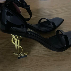 Saint Laurent svarta klackar  - Exklusiva svarta sandaler från Saint Laurent i lackat skinn med öppen tå och smala remmar. Klacken är i guld och formad som YSL-loggan, vilket ger en riktigt lyxig vibe. Perfekta för dig som vill sticka ut med en statement-sko.