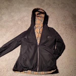 Burberry vindjacka med klassiskt rutigt foder - Svart vindjacka från Burberry med dragkedja och huva. Insidan har det ikoniska beige-rutiga Burberry-mönstret. Jackan har två fickor med knapp och justerbar nederkant. Perfekt för dig som vill ha en stilren och tidlös look med lyxig känsla. Priset är inte hugget I sten!