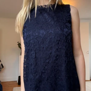 Mörkblå spetstopp utan ärm - Supersnygg mörkblå topp i spets med rak passform och ärmlös design. Toppen har en hög hals med fin spetskant och är helfodrad förutom längst ner där spetsen är genomskinlig. Perfekt för både vardag och finare tillfällen