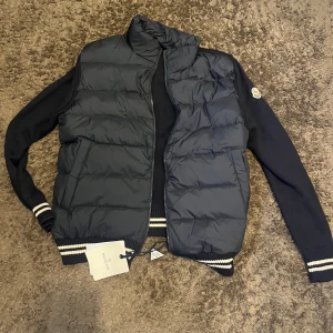 Mörkblå pufferjacka från Moncler - Snygg mörkblå pufferjacka från Moncler med stickade ärmar och quiltad front. Jackan har dragkedja, ribbade muddar med vita ränder och klassisk Moncler-logga på ärmen. Perfekt för höst och vinter när du vill hålla stilen och värmen.