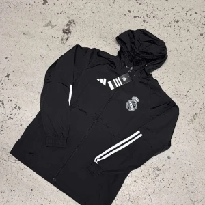 Svart vindjacka Real Madrid Adidas - Svart vindjacka från Adidas med Real Madrid-logga på bröstet. Jackan har huva, hel dragkedja och vita ränder längs ärmarna. Perfekt för dig som gillar sportig stil och vill visa ditt lagstöd.