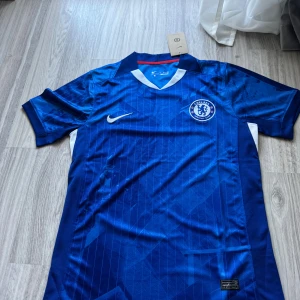 Chelsea FC hemmaställ 25/26 (Rabatt) - Chelsea FCs officiella blå matchtröja från Nike med klubbmärke på bröstet och vit Nike-logga. Tröjan har V-ringning med vit och röd detalj, korta ärmar och är tillverkad i Dri-FIT-material som andas. Snygga subtila mönster och vita/röda inslag på sidorna.