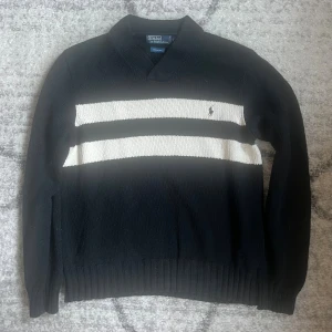 Svart stickad tröja Polo Ralph Lauren - Svart stickad tröja från Polo Ralph Lauren med två breda vita ränder över bröstet och broderad logga. Tröjan har v-ringad krage, ribbade muddar och är tillverkad i 100% bomull. Perfekt för en stilren och sportig look.