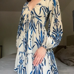 Blåvit blommig kortklänning H&M - Supersöt kortklänning från h&m med blått blommönster på vit botten. Klänningen har v-ringning, långa ärmar med resår vid handlederna och plisserat tyg som ger en flowy känsla. Perfekt för dig som gillar en luftig och trendig stil.