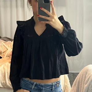 Svart blus med volang och v-ringning - Säljer en svart blus med v-ringning och volangdetaljer vid axlarna. Blusen har långa, lösa ärmar med lätt puff och är croppad i modellen. Tillverkad i ett mjukt bomullstyg som känns skönt mot huden. Perfekt för dig som gillar en trendig och avslappnad look.