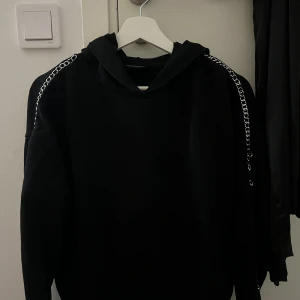 Svart hoodie med kedjedetaljer från Diesel - Svart hoodie från Diesel med coola silverfärgade kedjor längs axlarna. Tröjan har huva och är långärmad, perfekt för dig som gillar streetstyle och vill sticka ut lite extra. Materialet är mjukt och skönt, troligtvis bomull eller polyester.