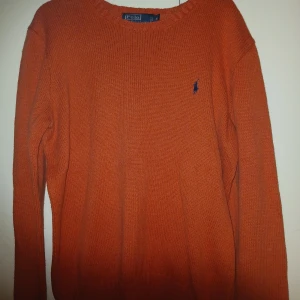 Orange stickad tröja från Polo Ralph Lauren - Säljer en snygg orange stickad tröja från Polo Ralph Lauren med klassisk blå logga broderad på bröstet. Tröjan har rund hals och långa ärmar, perfekt för dig som gillar stilrena och färgstarka plagg.