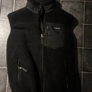 Svart fleeceväst från Patagonia - Svart fleeceväst från Patagonia med hög krage och flera praktiska fickor med dragkedja. Västen har ett mjukt, fluffigt material och detaljer i syntet på fickorna och axlarna. Perfekt för lager-på-lager och outdoor-vibes.