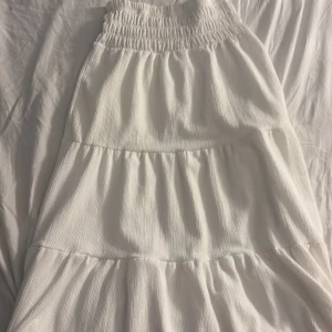 Vit smockad sommarklänning från Gina Tricot - Superfin vit klänning från Gina Tricot med smock upptill och volangiga lager. Klänningen har en luftig känsla och är perfekt för varma dagar. Materialet är mjukt och lätt, och modellen är flowy med snygg struktur.