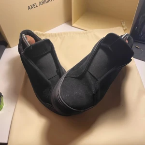 Svarta sneakers från Axel Arigato - Snygga svarta sneakers från Axel Arigato i mocka. De har en stilren design utan snörning. Skorna är i bra skick men mockan är lite förstörd på högra skon. Lite gamla bilder nu är den väl använda. Den funkar men hål i sulan och mockan är o räddbar