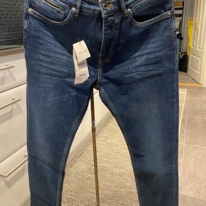 Slim fit blå jeans med snygga detaljer - Slim fit jeans i klassisk blå tvätt med snygga kontrastsömmar och femficksdesign. Byxorna har raka ben och är tillverkade i jeansmaterial. Passar dig som gillar en stilren look med lite smalare passform. Märkt med storlek 40 och har etikett kvar.